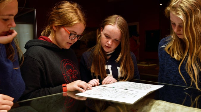 Four young teenage girls look at a music book in a glass case. The background is dark. They have a sheet of paper information in front of them.